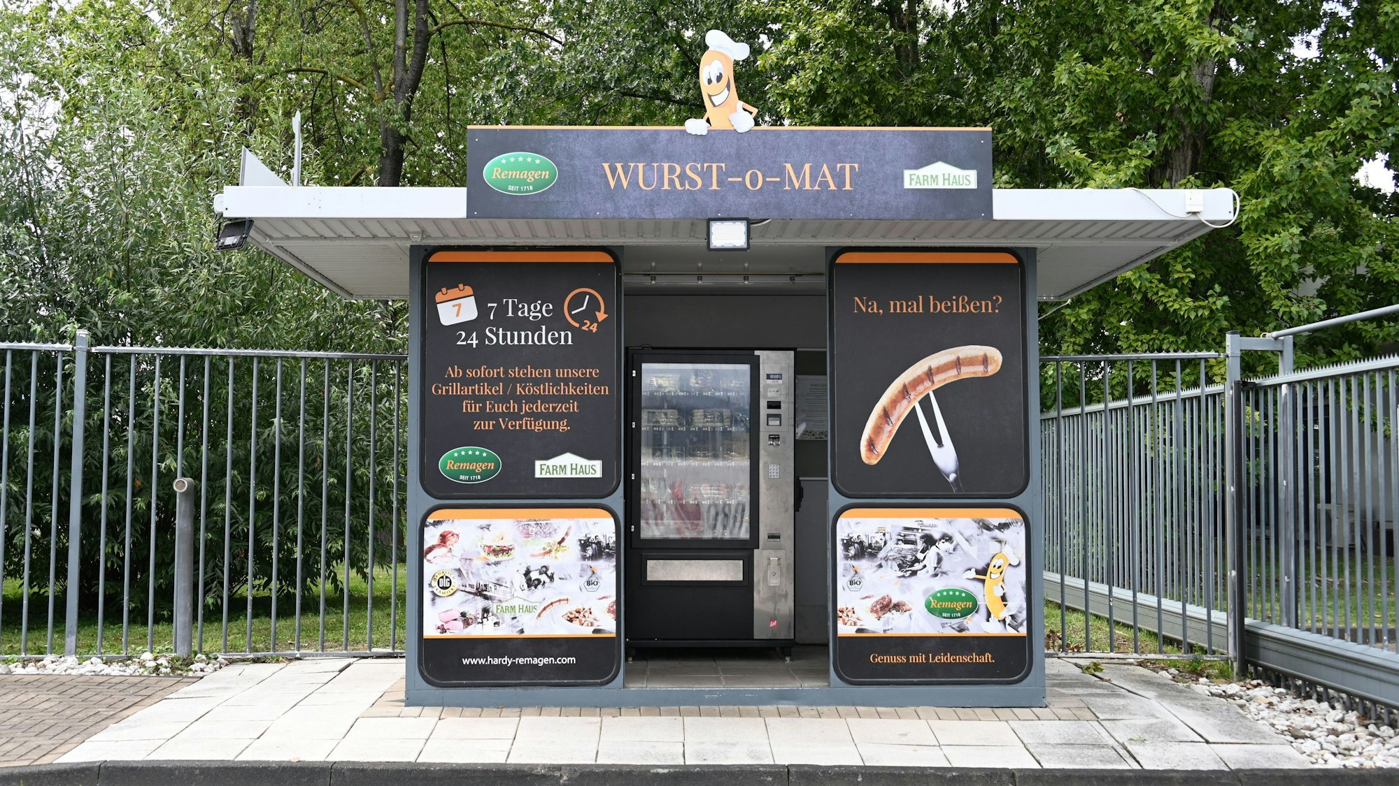 Ein Häuschen mit der Aufschrift „Wurst-o-Mat“. Rechts und links sind Schilder mit der Aufschrift „7 Tage, 24 Stunden“ und „Na, mal beißen?“, dazu das Bild einer Wurst auf einer Gabel. Durch den Eingang kann man den Automaten erkennen.
