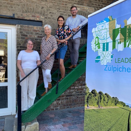 Auf einer grünen Treppe vor einer Wand stehen: die Weilerswister Bürgermeisterin Anna-Katharina Horst (v.l.), als Projektträgerin Marietta Thien, Projektmitarbeiterin Sarah Rodewald und Regionalmanager des LAG Zülpicher Börde, Sebastian Duif.
