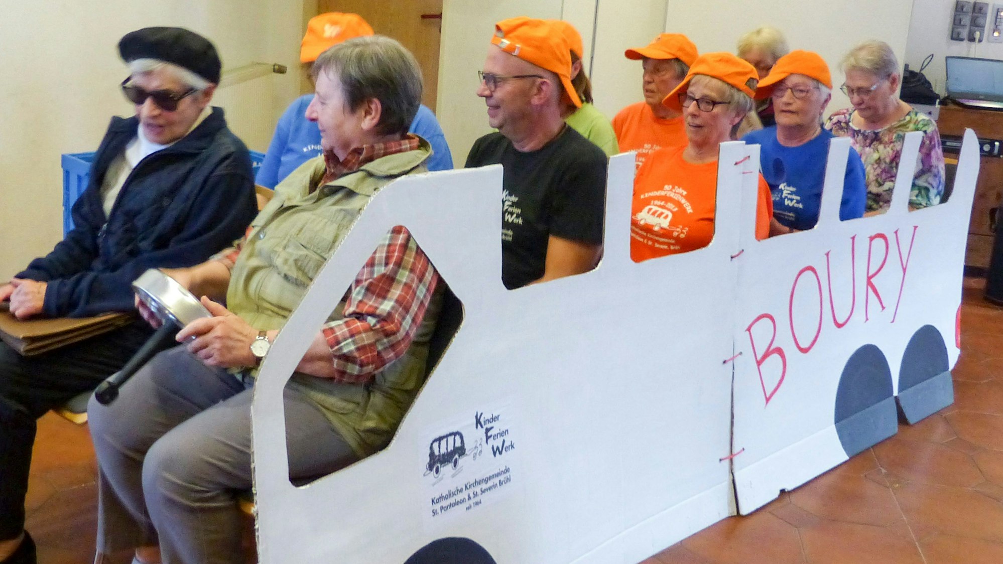 Das Bild zeigt zehn ehemalige Betreuer, die in einem Bus aus Pappe sitzen. Sie tragen zum großen Teil orange Kappen.