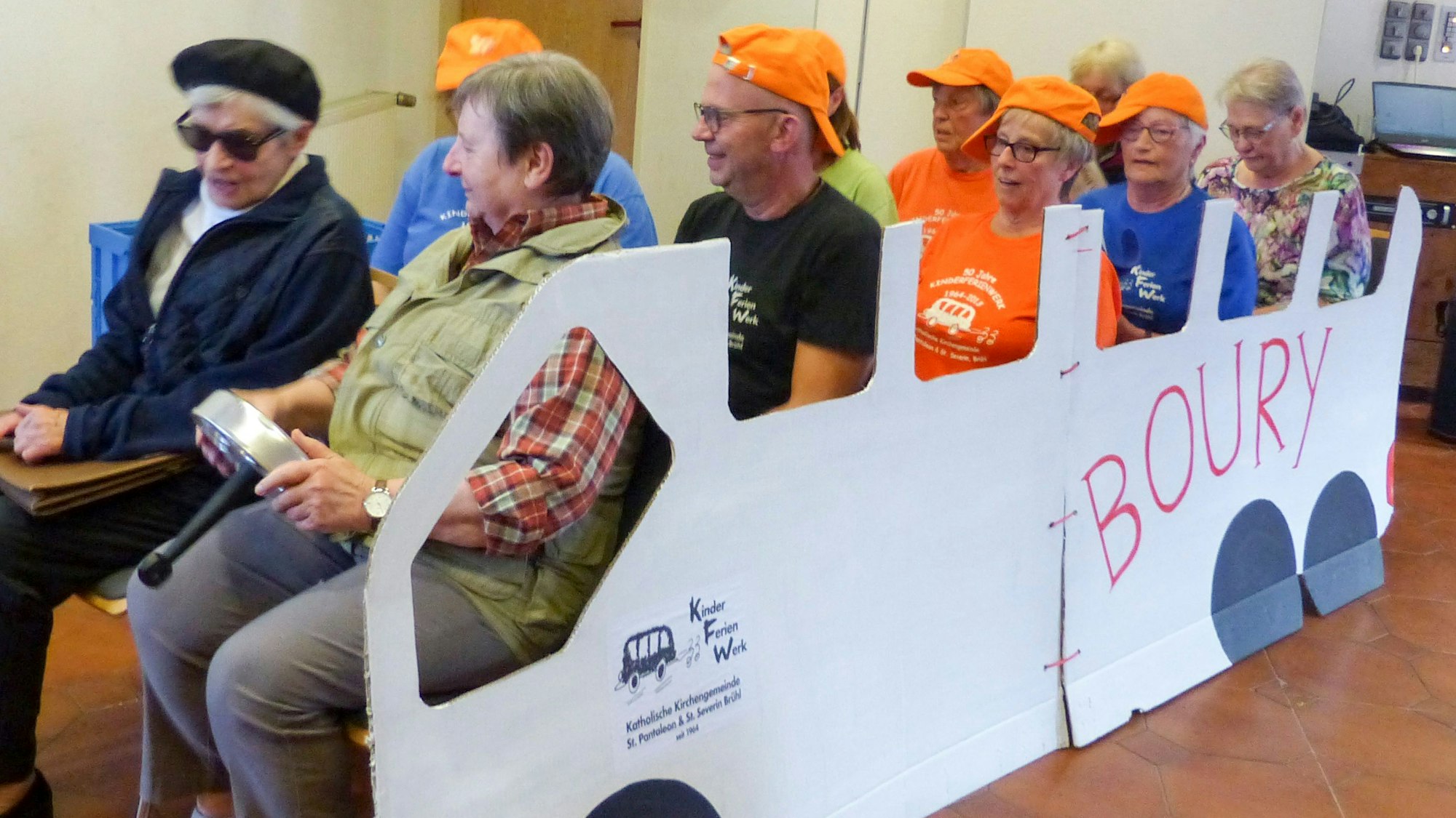 Das Bild zeigt zehn ehemalige Betreuer, die in einem Bus aus Pappe sitzen. Sie tragen zum großen Teil orange Kappen.