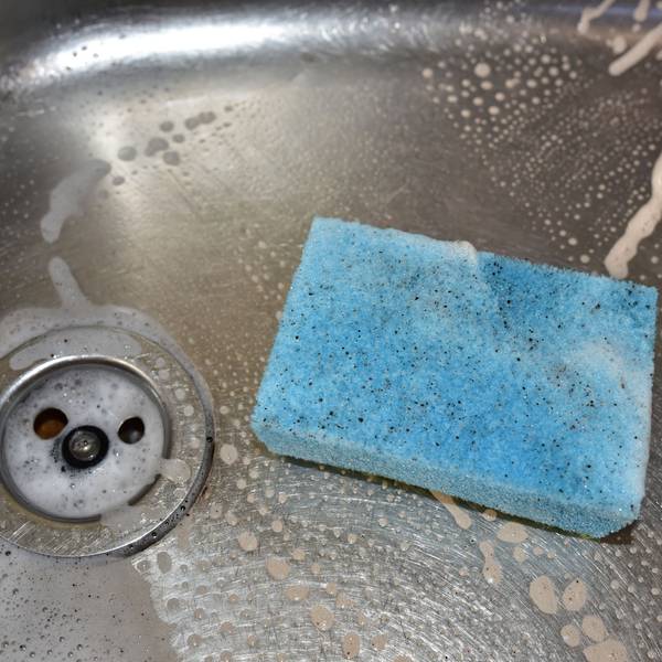 Ein dreckiger, hellblauer Spülschwamm liegt in einer Spüle, in der außerdem Rückstände von Spülmittel zu sehen sind.