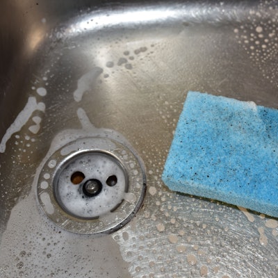 Ein dreckiger, hellblauer Spülschwamm liegt in einer Spüle, in der außerdem Rückstände von Spülmittel zu sehen sind.