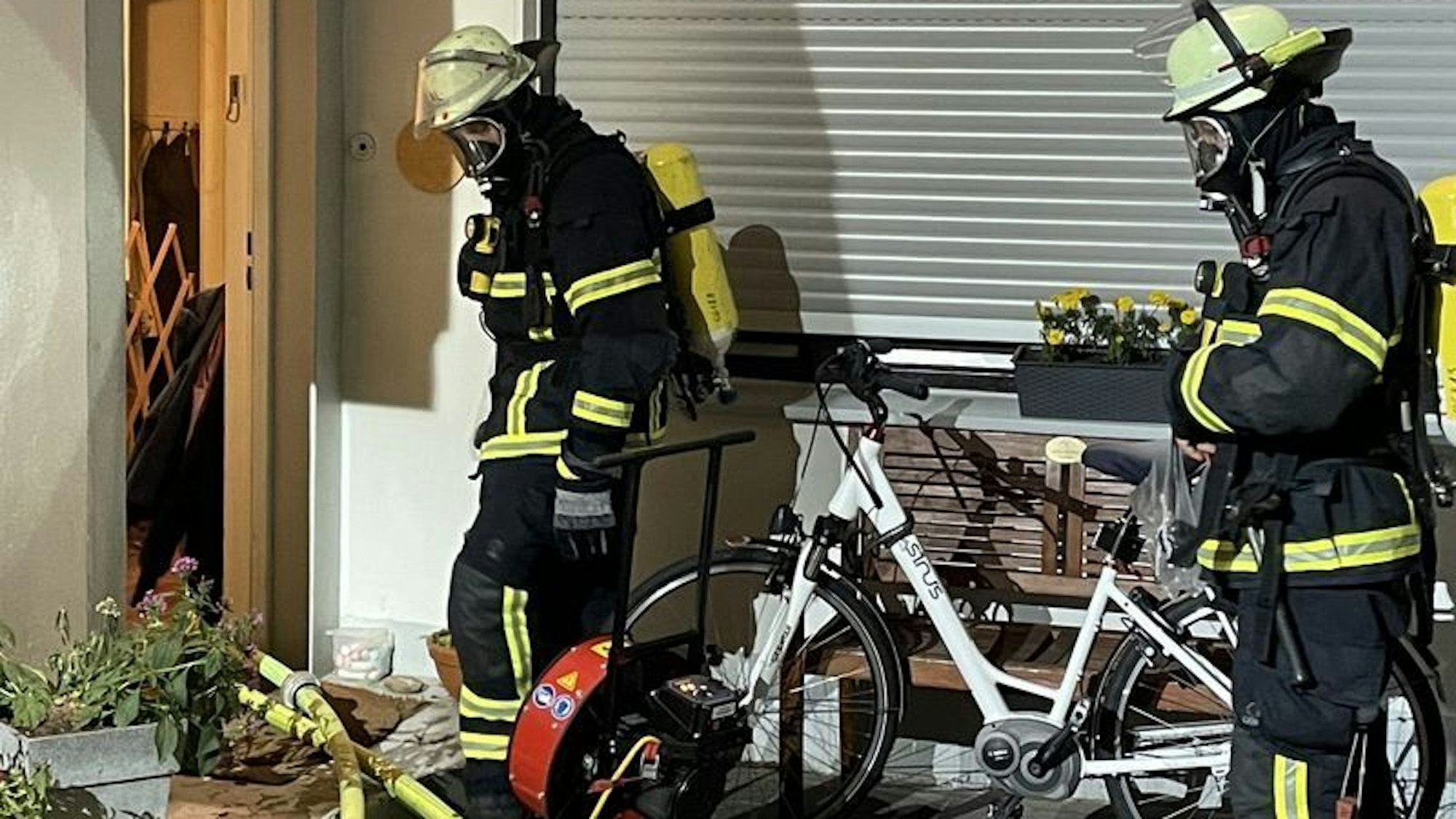 Zwei Feuerwehrleute bei einem Brand in Königswinter. Sie stehen in voller Ausrüstung am Hauseingang neben einem Lüfter, der den Rauch aus dem Haus bläst.