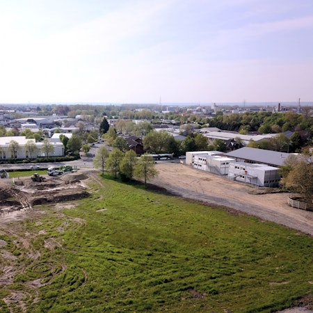 Aufsicht von oben auf ein gerodetes Areal mit zerfahrener Wiese, an deren Rand Baustellenfahrzeuge stehen. Bebauung ist sichtbar.
