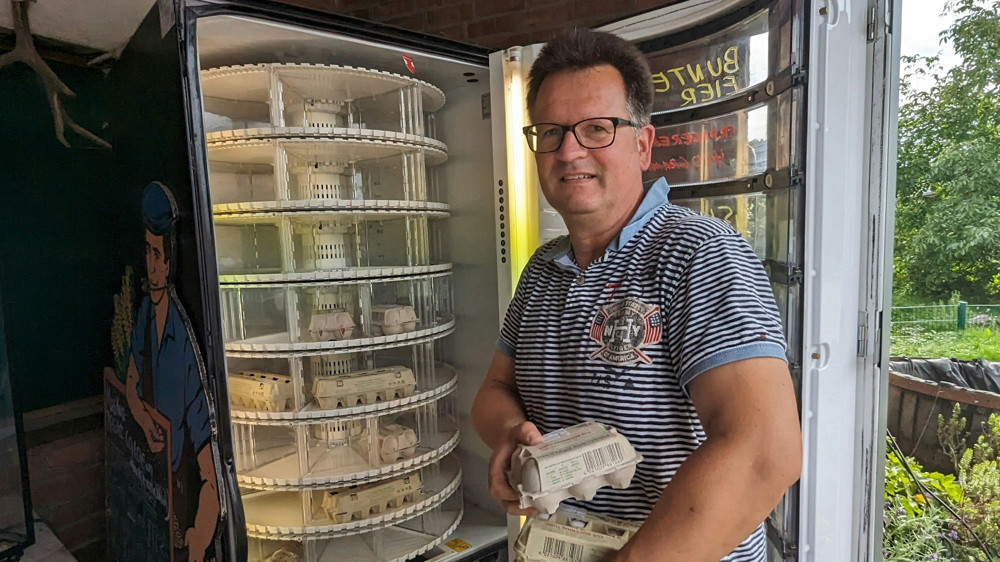 Der Landwirt hält einen Stapel Eierkartons im Arm. Der Automat vor ihm ist geöffnet. Einige Eierkartons sind bereits im Automaten.