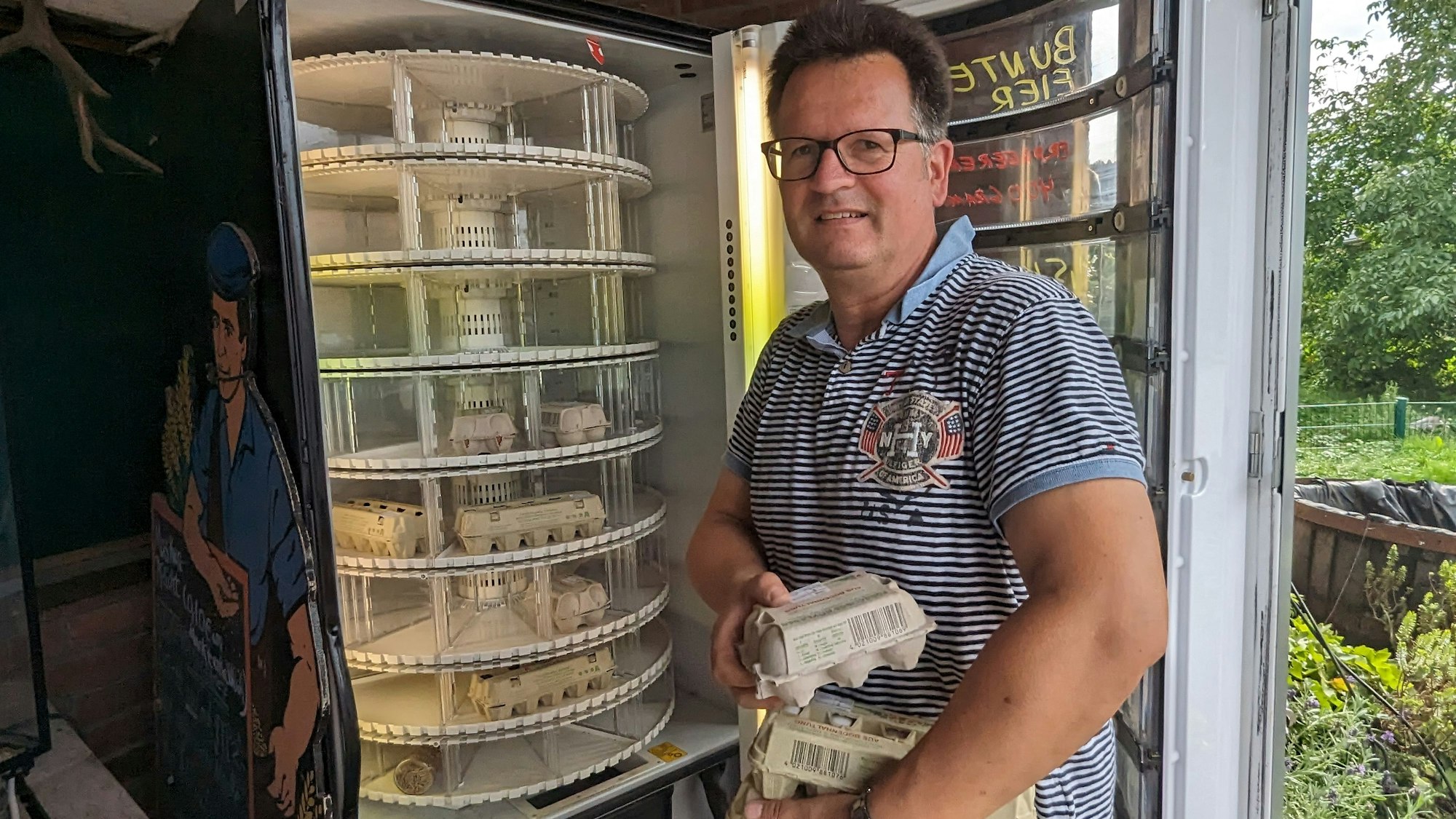 Der Landwirt hält einen Stapel Eierkartons im Arm. Der Automat vor ihm ist geöffnet. Einige Eierkartons sind bereits im Automaten.