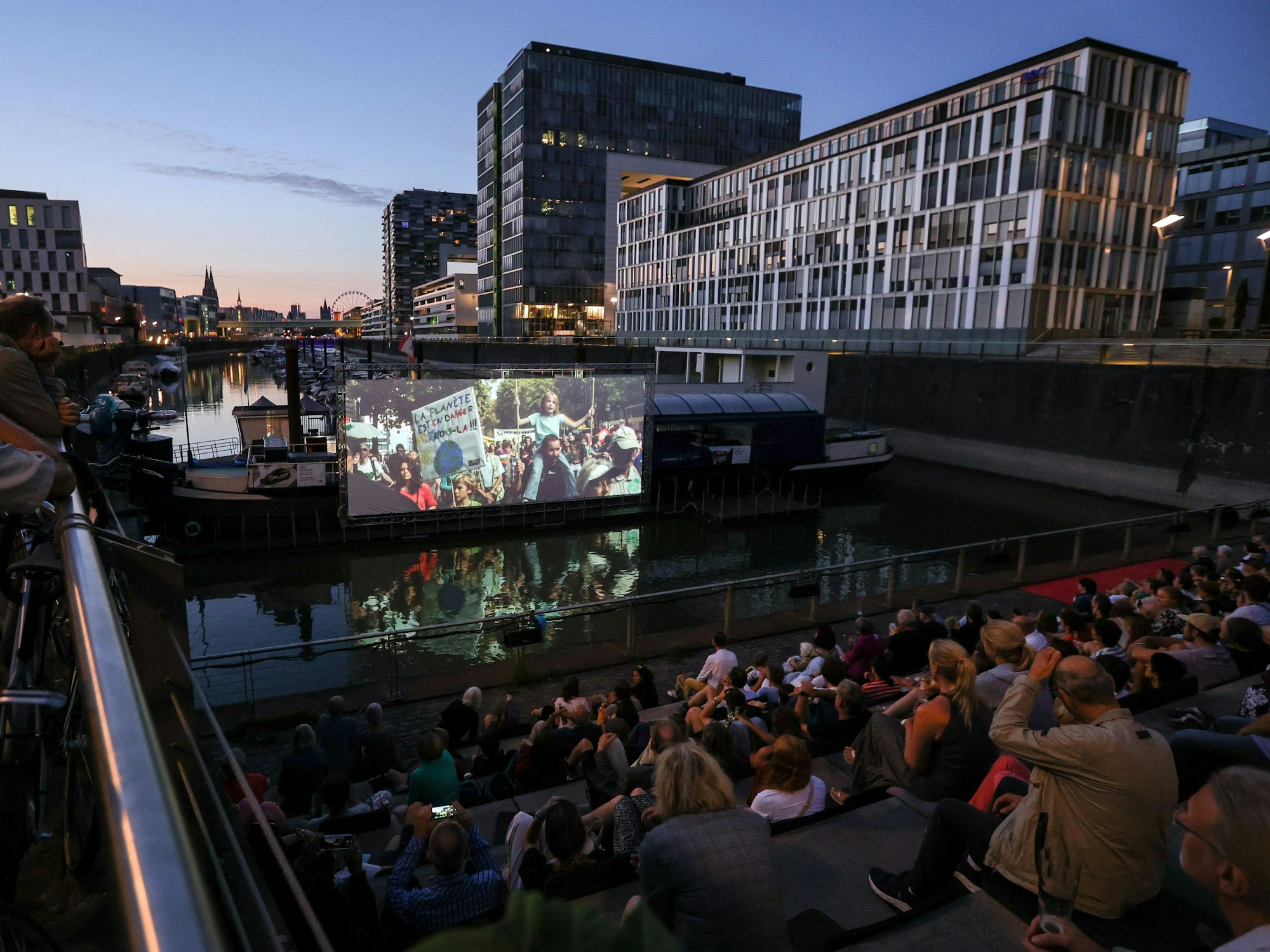 Kinoabend am Rheinauhafen: Menschen sitzen vor einer Leinwand auf dem Wasser des Yachthafens in Köln. Im Hintergrund ist der Kölner Dom in der Abenddämmerung zu sehen.