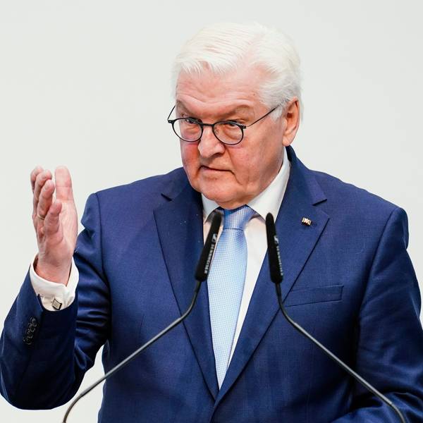 Bundespräsident Frank-Walter Steinmeier steht vor Mikrofonen und hebt eine Hand vor einem weißen Hintergrund.