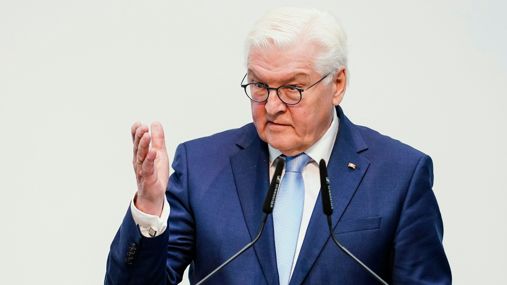 Bundespräsident Frank-Walter Steinmeier steht vor Mikrofonen und hebt eine Hand vor einem weißen Hintergrund.