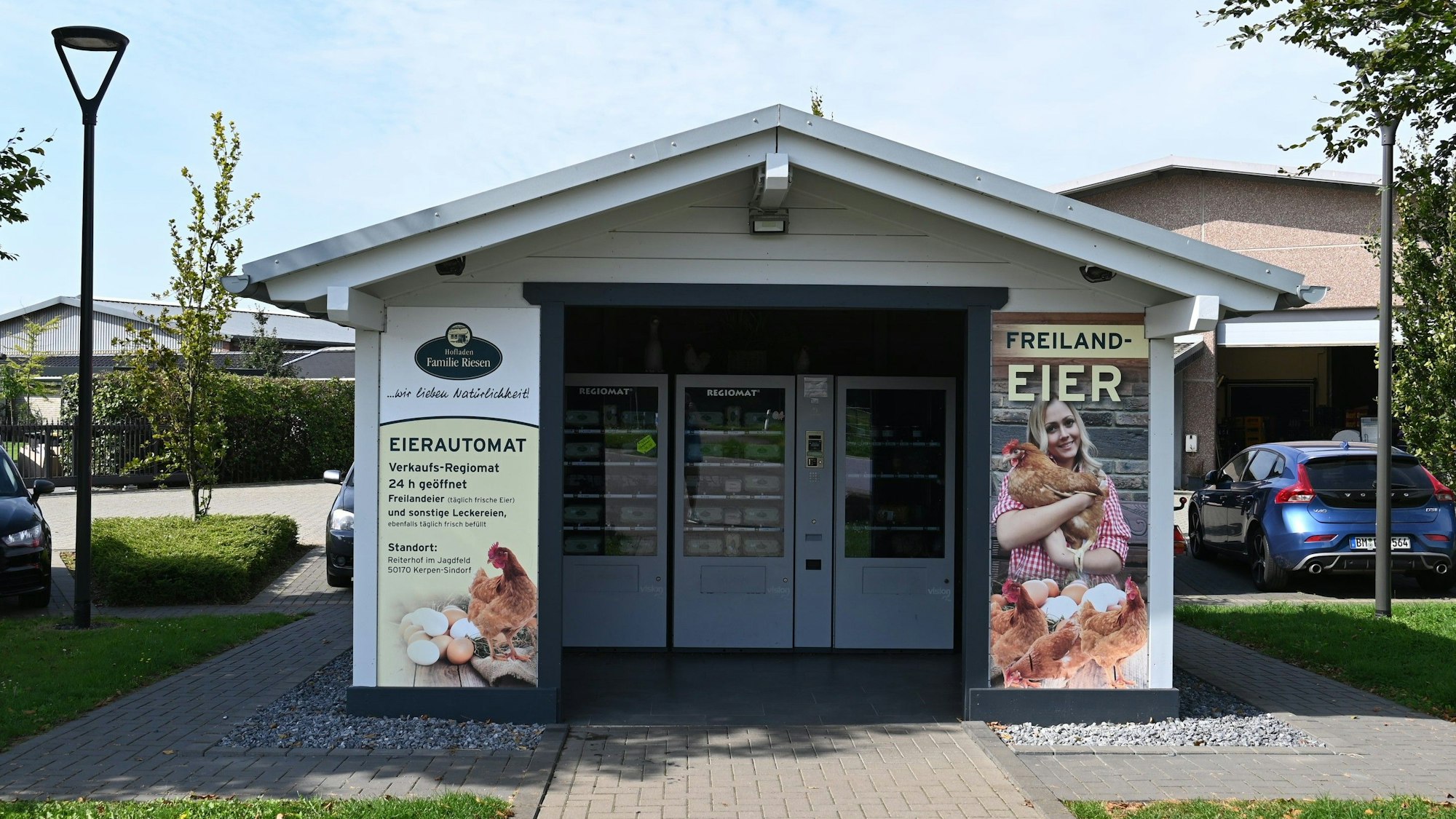 Ein Häuschen. Rechts die Aufschrift „Freiland-Eier“ und das Bild einer Frau mit einem Huhn im Arm. Links das Wort „Eierautomat“ und weitere Informationen. Durch den Eingang kann man mehrere Automaten sehen.