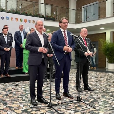 Bundeskanzler Olaf Scholz steht mit zwei weiteren Männern vor mehreren Mikrofonen, im Hintergrund haben sich Bürgermeisterinnen und Bürgermeister aufgestellt.