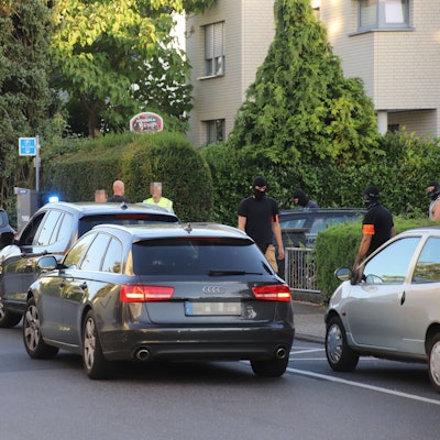 Mehrere Autos stehen auf einer Straße mit Mehrfamilienhäusern, Männer mit Sturmhauben stehen daneben.