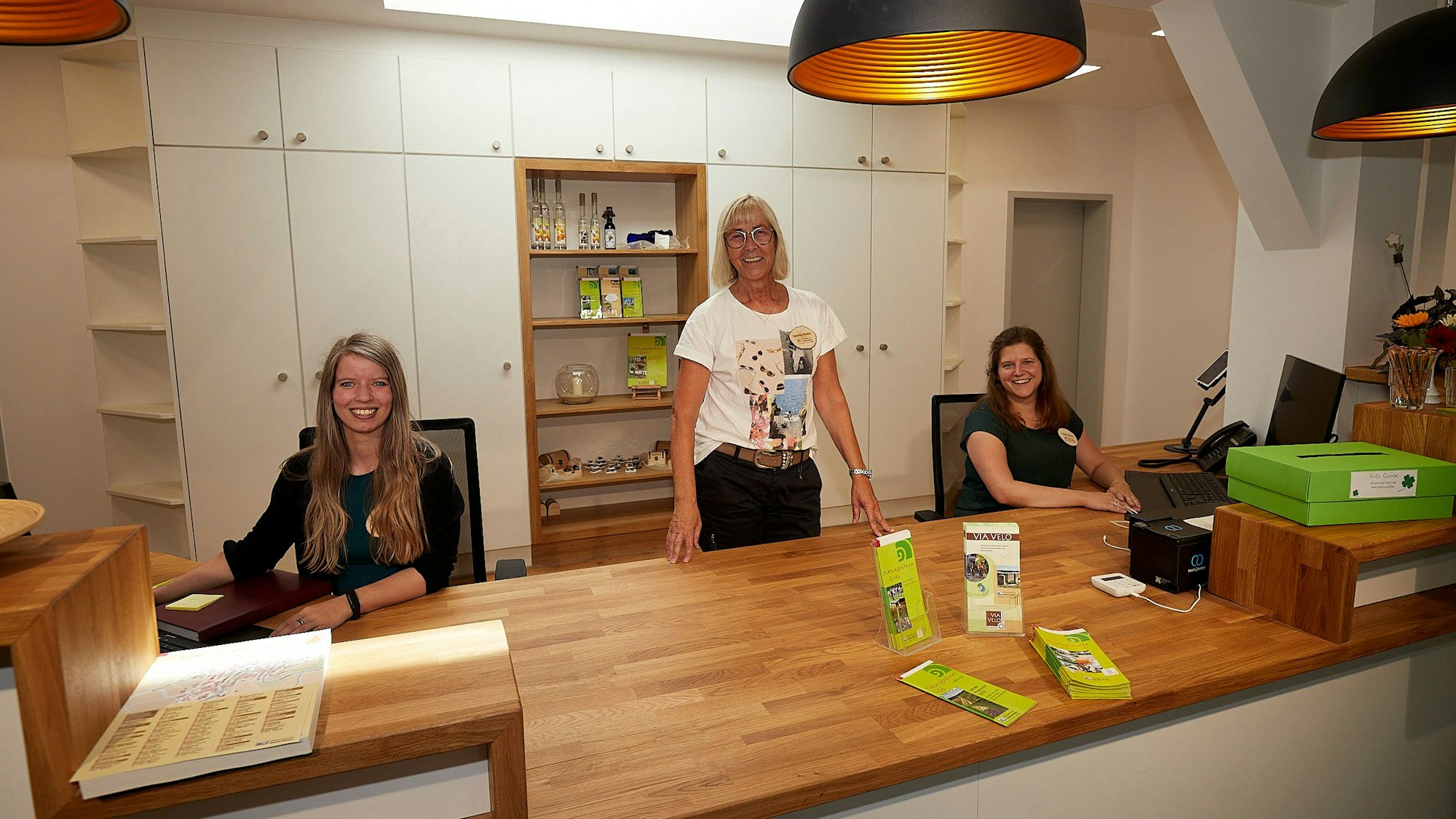 Drei Frauen, Sophie Bey, Veronika Hoeller und Andrea Düren, sind an ihrem Arbeitsplatz hinter der Theke der Tourist-Information in Nettersheim, die in weiß und mit Holz modern gestaltet ist.