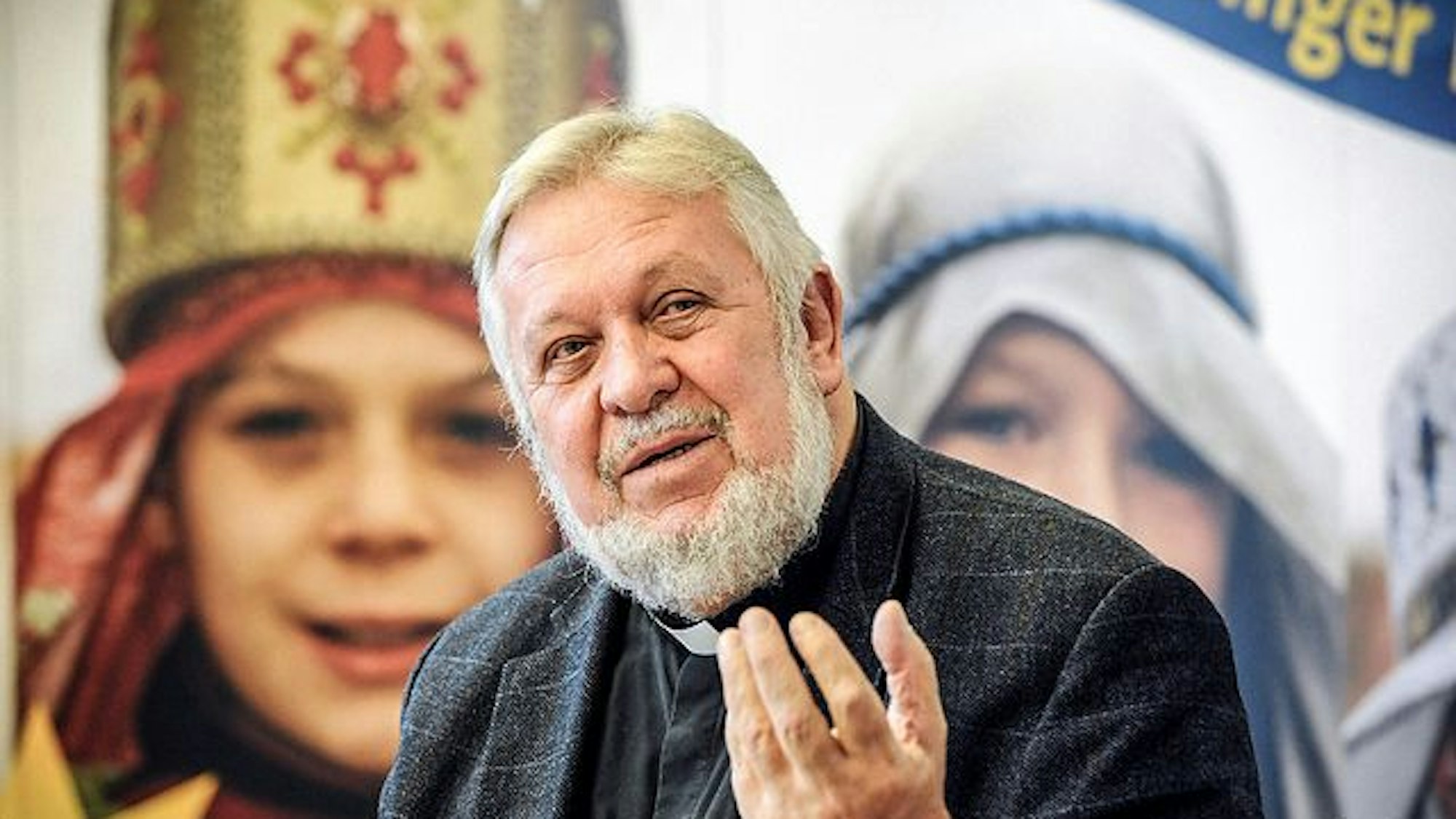 Alter Mann mit Bart und Priesterkragen vor einem Bild mit Sternsingern.