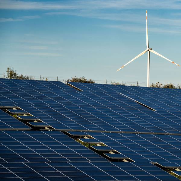 Ein Windrad steht auf einem Hügel oberhalb einer großflächigen Solaranlage auf einem Feld.