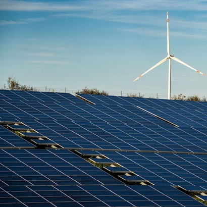 Ein Windrad steht auf einem Hügel oberhalb einer großflächigen Solaranlage auf einem Feld.