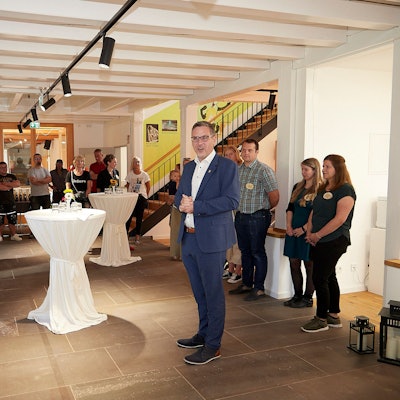 In einem großen Raum stehen Stehtische mit weißen Hussen. In der Mitte steht Nettersheims Bürgermeister Norbert Crump, an den Wänden zahlreiche andere Menschen.  Sie feiern die Wiedereröffnung des Naturzentrums.