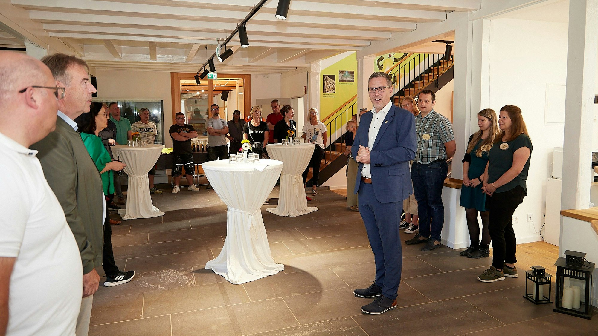 In einem großen Raum stehen Stehtische mit weißen Hussen. In der Mitte steht Nettersheims Bürgermeister Norbert Crump, an den Wänden zahlreiche andere Menschen.  Sie feiern die Wiedereröffnung des Naturzentrums.