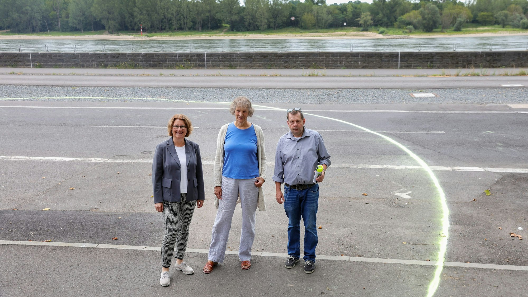 Zwei Frauen und ein Mann mittleren Alters stehen auf einer Straße vor dem Rheinufer