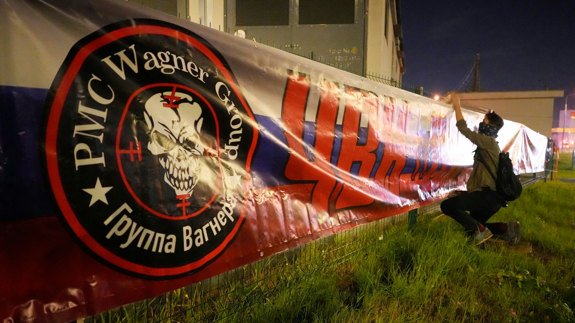 Ein Mann hängt ein Banner in Sankt Petersburg mit dem Logo der Wagner-Gruppe auf, der Schriftzug lautet: Wir stehen zusammen.