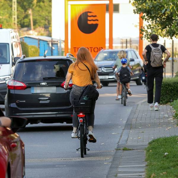 Autos und Fahrräder auf einer engen Straße vor dem Schulcampus in Vogelsang.