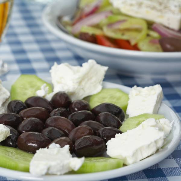Teller mit Gurken, Oliven und Feta und einem Glas Retsina auf einer blau-weißen Tischdecke