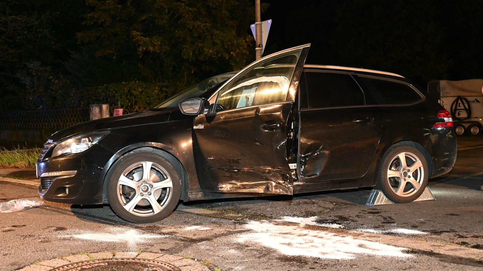 Ein dunkles Auto steht mit eingedellter Seite und offener Fahrertür an der Unfallstelle.