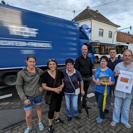 Eine Gruppe von Männern und Frauen steht an einer Straße, im Hintergrund ein Lkw und ein Bus.