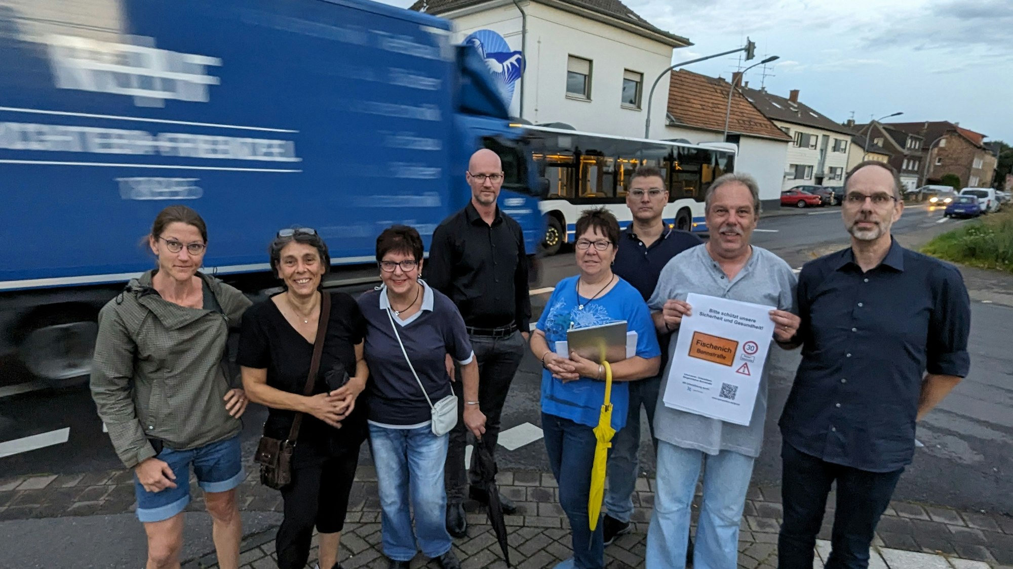Eine Gruppe von Männern und Frauen steht an einer Straße, im Hintergrund ein Lkw und ein Bus.