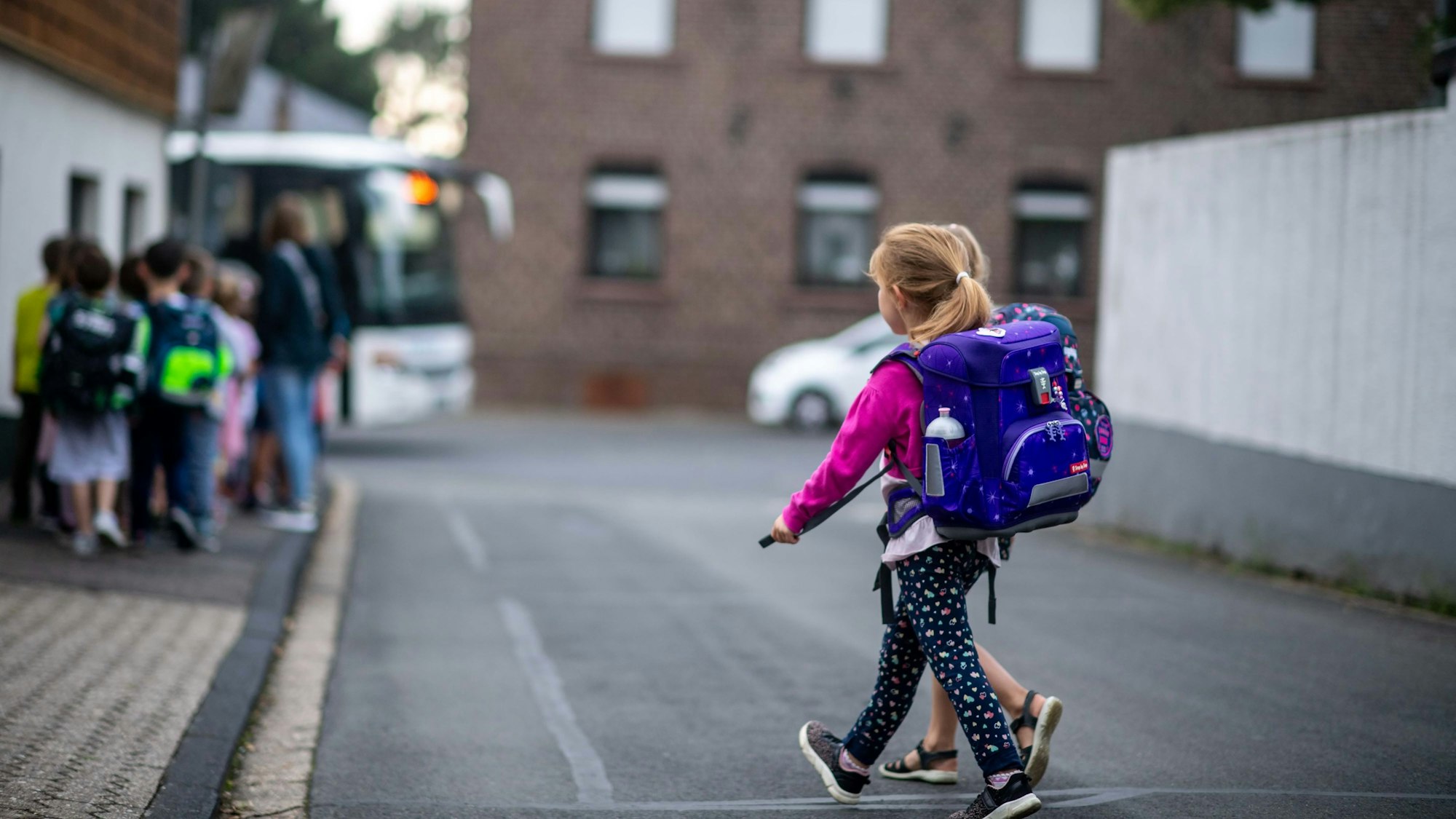 Das Bild zwei Grundschülerinnen, wie sie eine Straße überqueren. Im Hintergrund geht eine weitere Gruppe in Richtung Bus.
