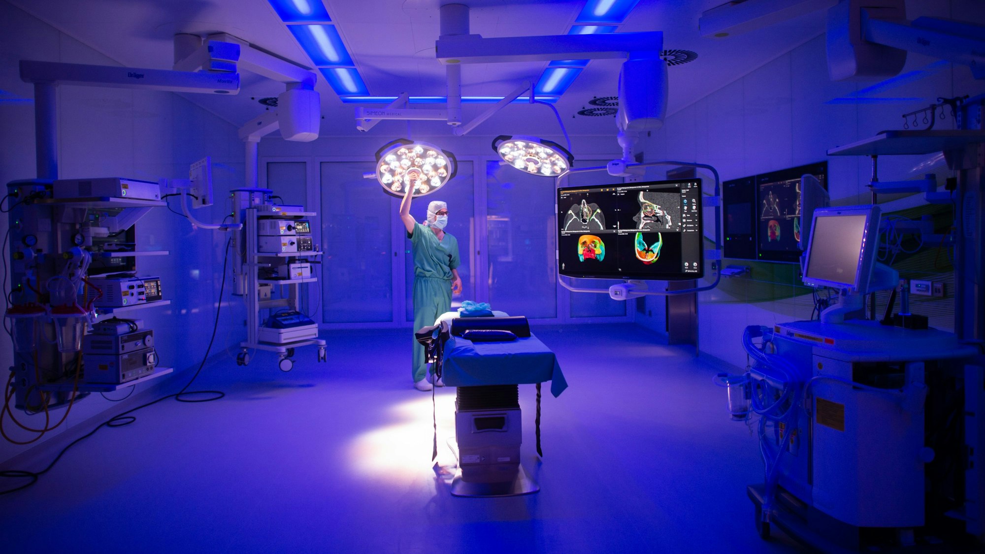 Blick in einen Operationssaal mit vielen technischen Geräten, während blaues Licht den Raum ausleuchtet und ein Mann die OP-Lampe einstellt.