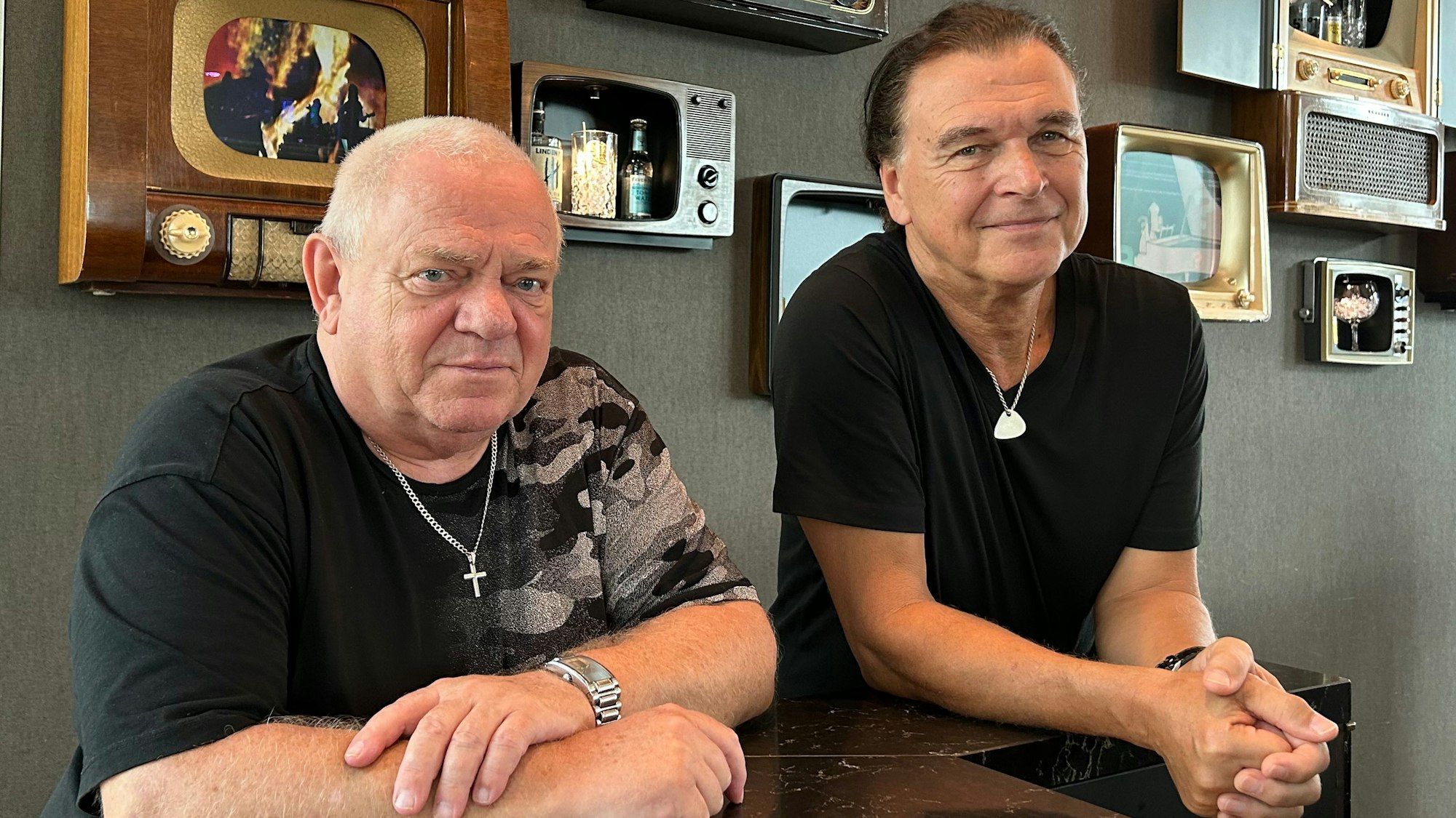 Udo Dirkschneider und Peter Baltes lehnen an einem Tresen, im Hintergrund sind historische Fernseher und Radiogeräte zu sehen.