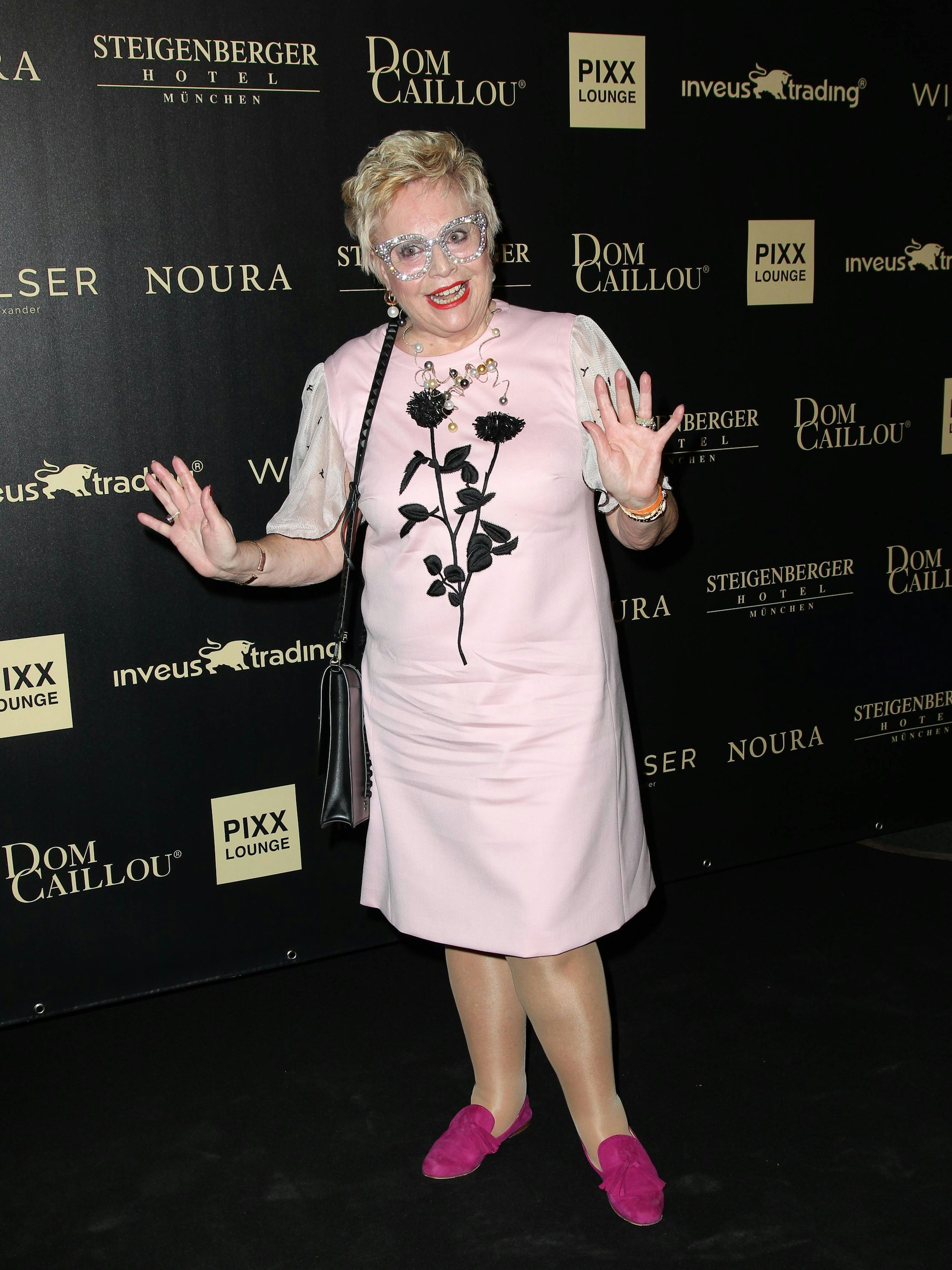 Maritta Emser steht in rosa Schuhen, einem hellrosa Kleid mit Blumenaufdruck und mit einer Glitzerbrille auf einem Event-Teppich in München.