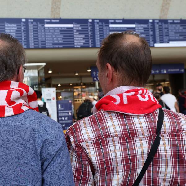 Zwei FC Fans mit rot-weißen Schals vor der Anzeigetafel am Kölner Hauptbahnhof.
