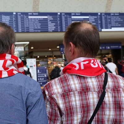 Zwei FC Fans mit rot-weißen Schals vor der Anzeigetafel am Kölner Hauptbahnhof.