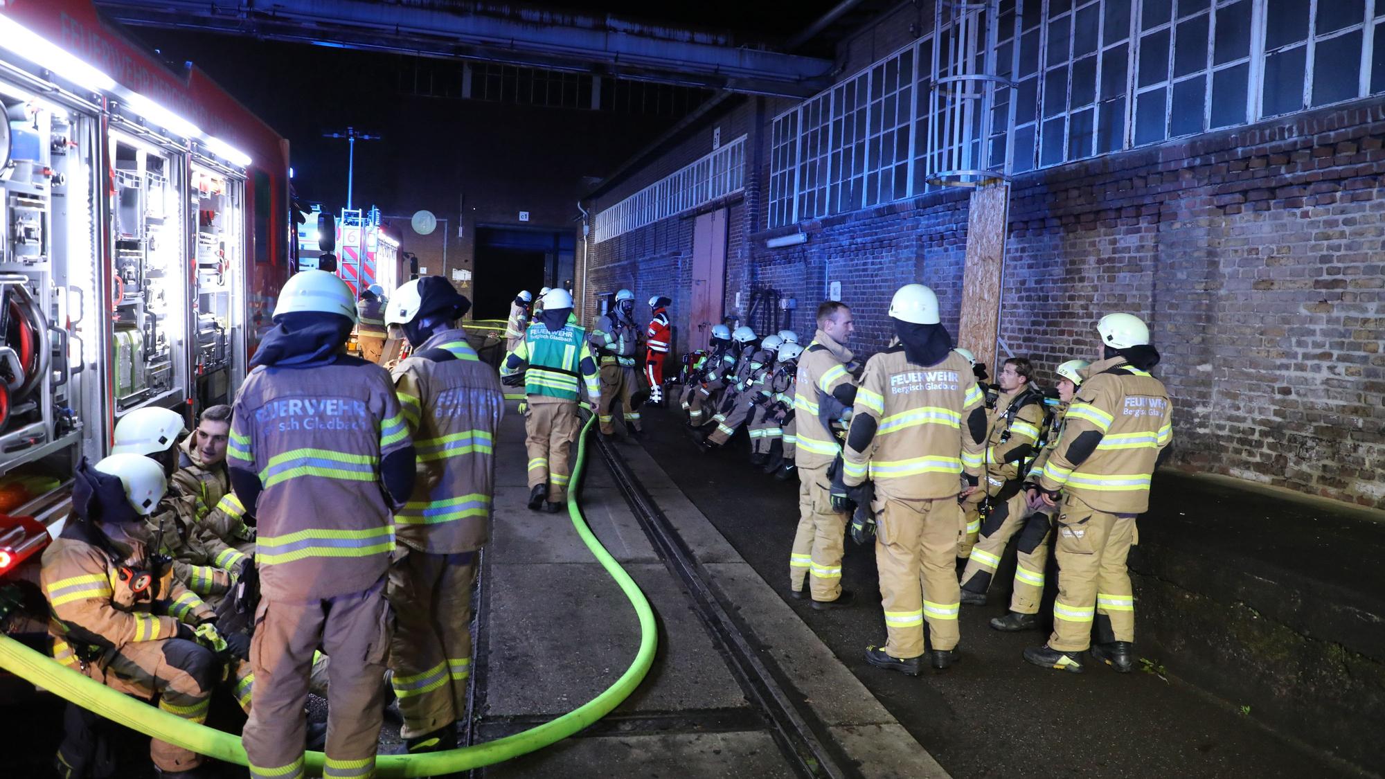 Feuerwehrleute arbeiten zwischen Feuerwehrfahrzeugen und einer Halle auf dem Zanders-Gelände.