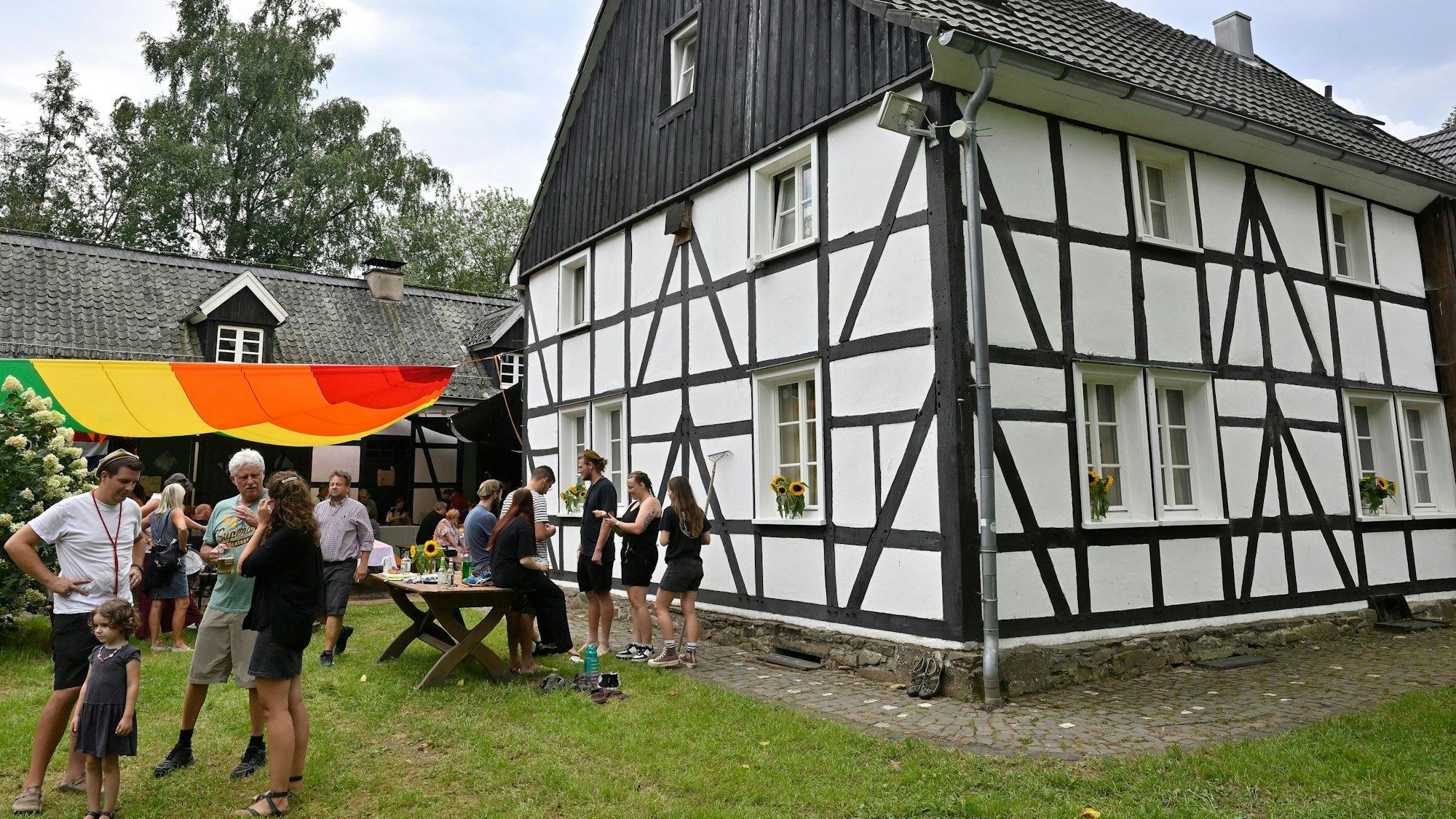Ein Fachwerkhaus, davor Leute unter einem bunten Sonnensegel, die feiern und sich unterhalten.
