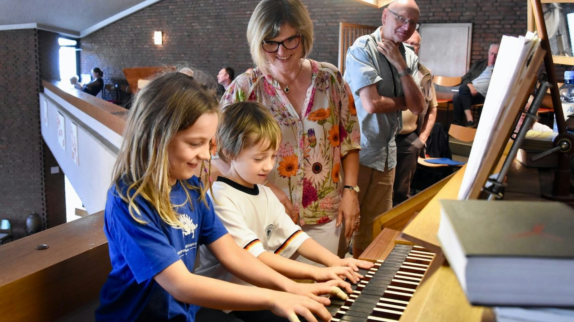 Ein Junge und ein Mädchen spielen auf einer Orgel. Eine Frau in bunter Bluse schaut, im Hintergrund steht ein weiterer Zuschauer.