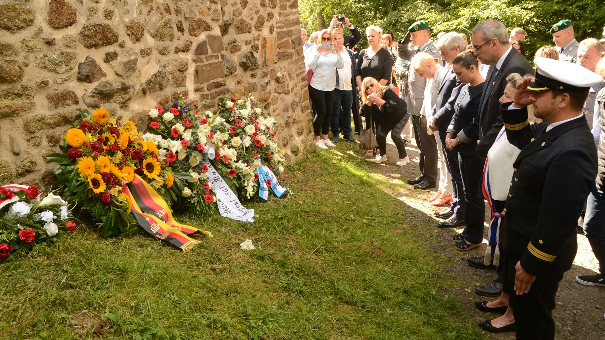 An einer alten Mauer liegen Trauerkränze. Im Vordergrund salutiert ein Marinesoldat, Männer und Frauen verneigen sich.
