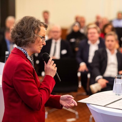 Das Bild zeigt Ministerin Ina Scharrenbach mit einem Mikrofon in der Hand, wie sie zum Publikum spricht.