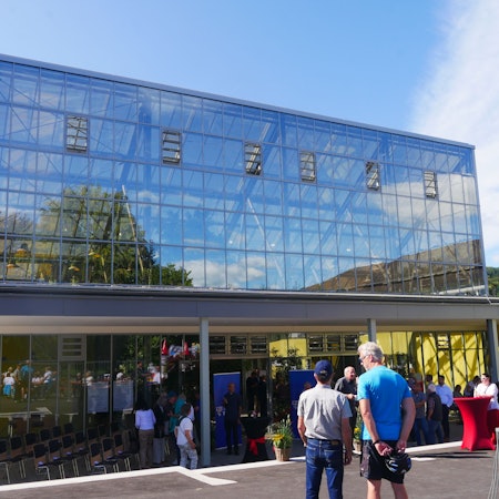 Vor einem großen gläsernen Gebäude stehen Besucher. In den Glasscheiben spiegeln sich blauer Himmel, Bäume und Wolken.