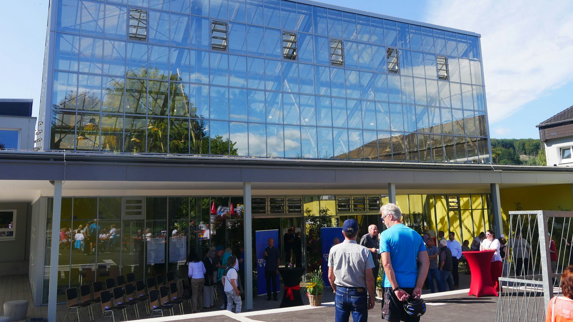 Vor einem großen gläsernen Gebäude stehen Besucher. In den Glasscheiben spiegeln sich blauer Himmel, Bäume und Wolken.