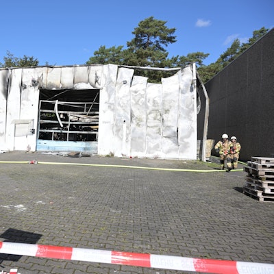 Absperrband prangt vor einer ausgebrannten Halle, Feuerwehrleute gehen auf einem Kontrollgang um die Halle.