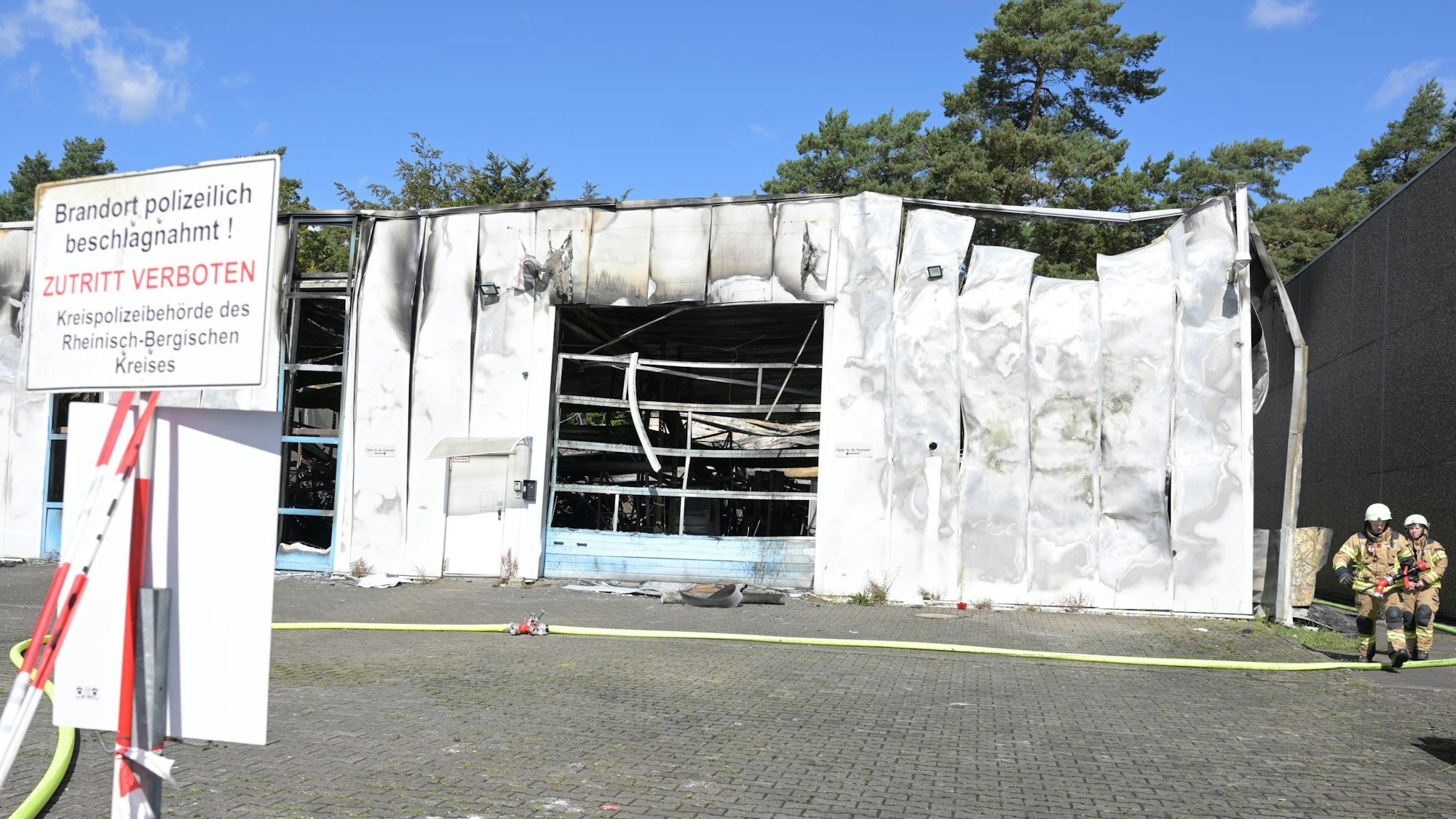 Absperrband prangt vor einer ausgebrannten Halle, Feuerwehrleute gehen auf einem Kontrollgang um die Halle.
