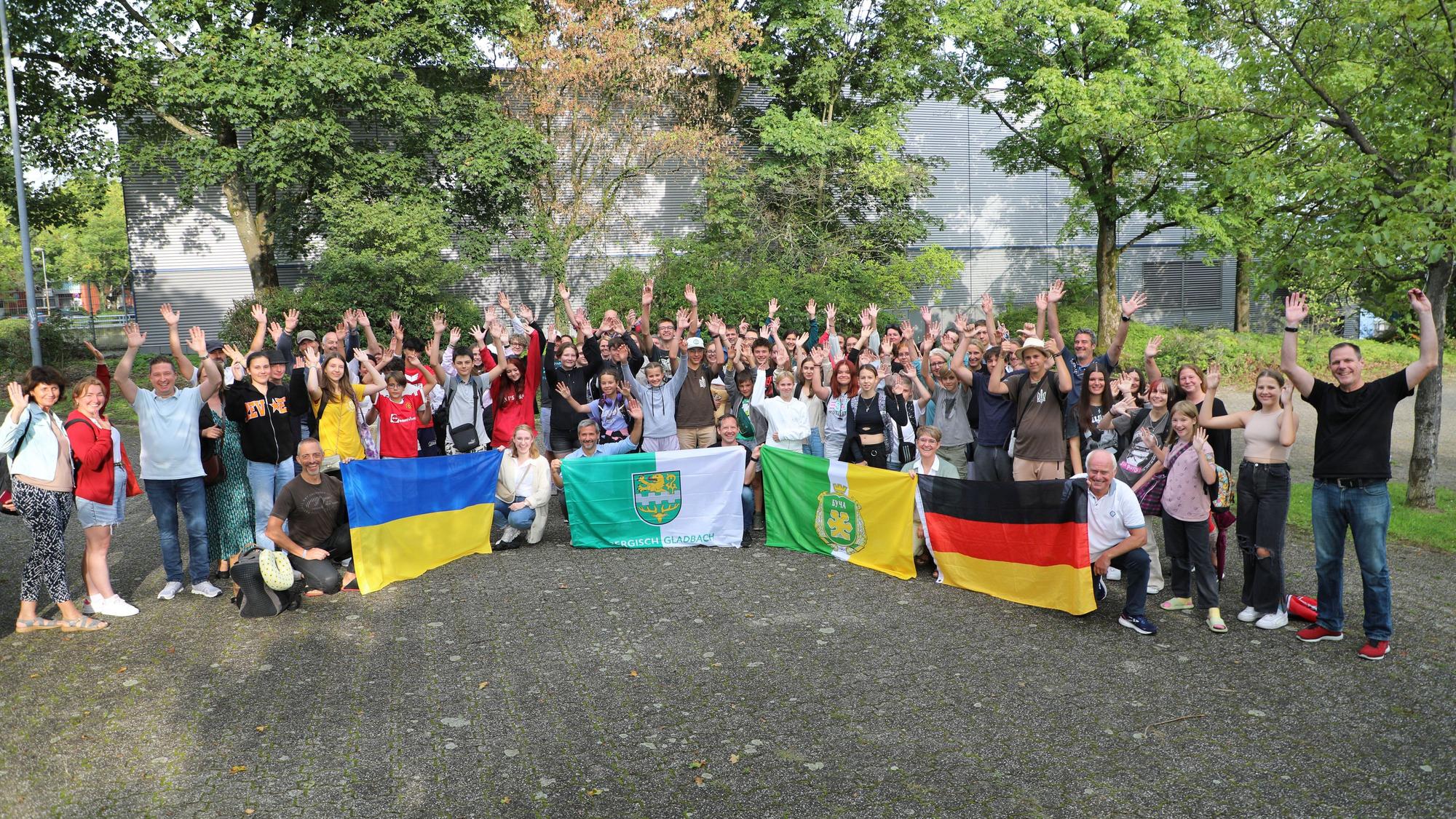 Schülerinnen und Schüler, Lehrerinnen und Lehrer und Eltern und andere Menschen stehen mit Fahnen von Ukraine, Deutschland, Butscha und Bergisch Gladbach auf dem Schulhof der Integrierten Gesamtschule Paffrath und jubeln.