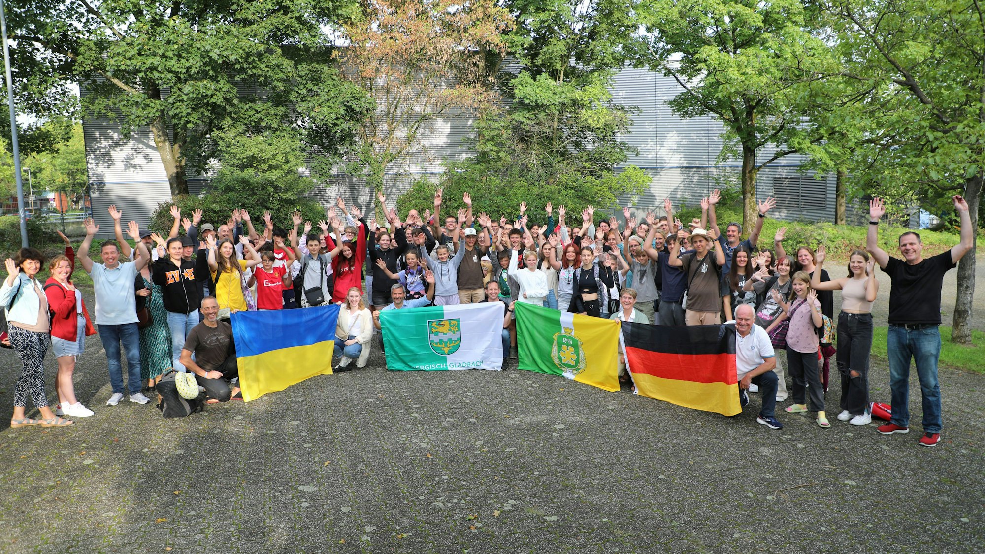 Schülerinnen und Schüler, Lehrerinnen und Lehrer und Eltern und andere Menschen stehen mit Fahnen von Ukraine, Deutschland, Butscha und Bergisch Gladbach auf dem Schulhof der Integrierten Gesamtschule Paffrath und jubeln.