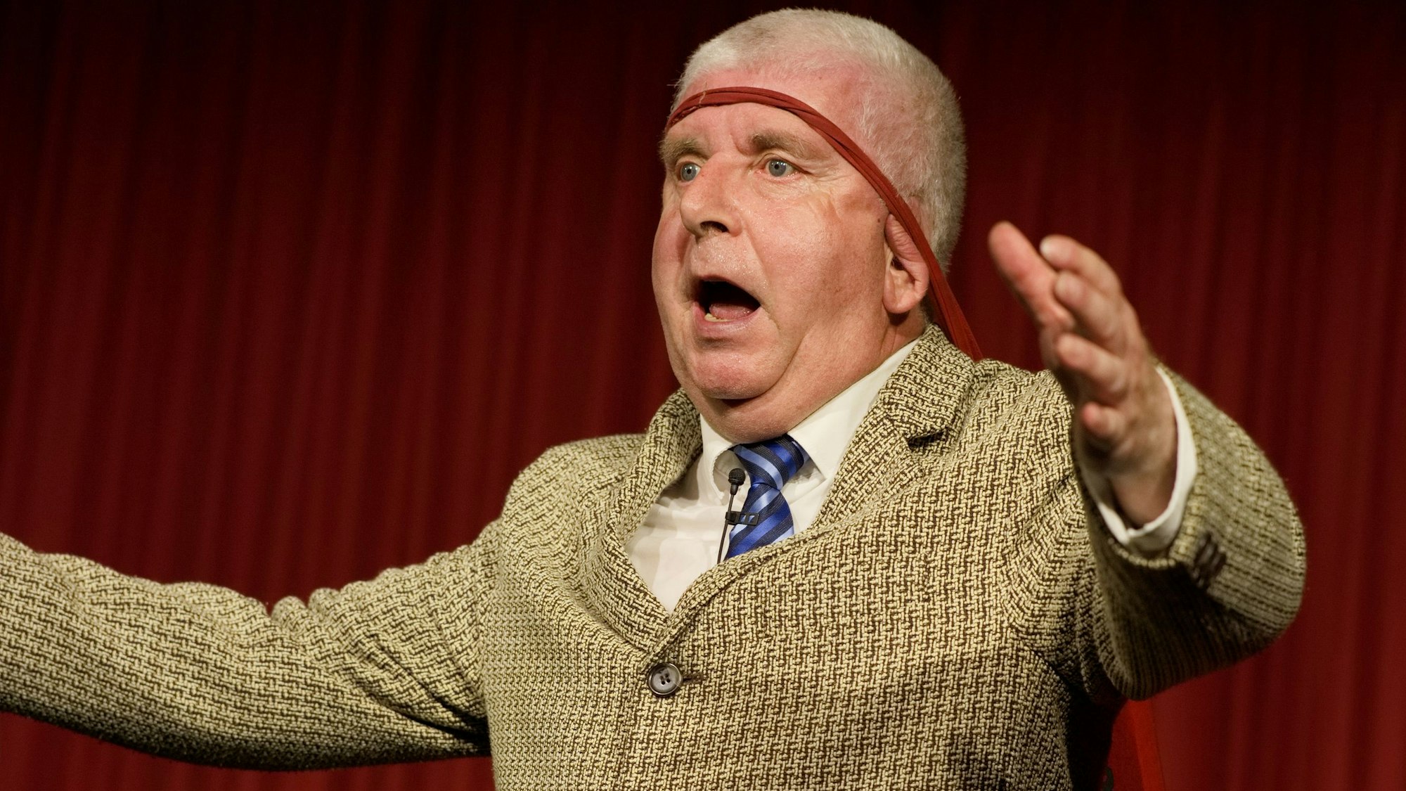 Das Bild zeigt den Kabarettisten Erwin Grosche, wie er auf der Bühne seine Arme ausbreitet.