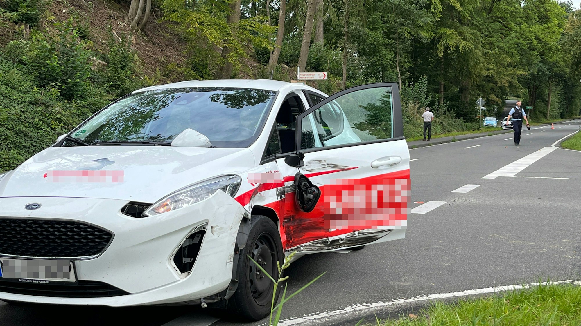 Auf der Straße steht ein weiß-rotes Auto, die Fahrerseite ist eingedrückt.