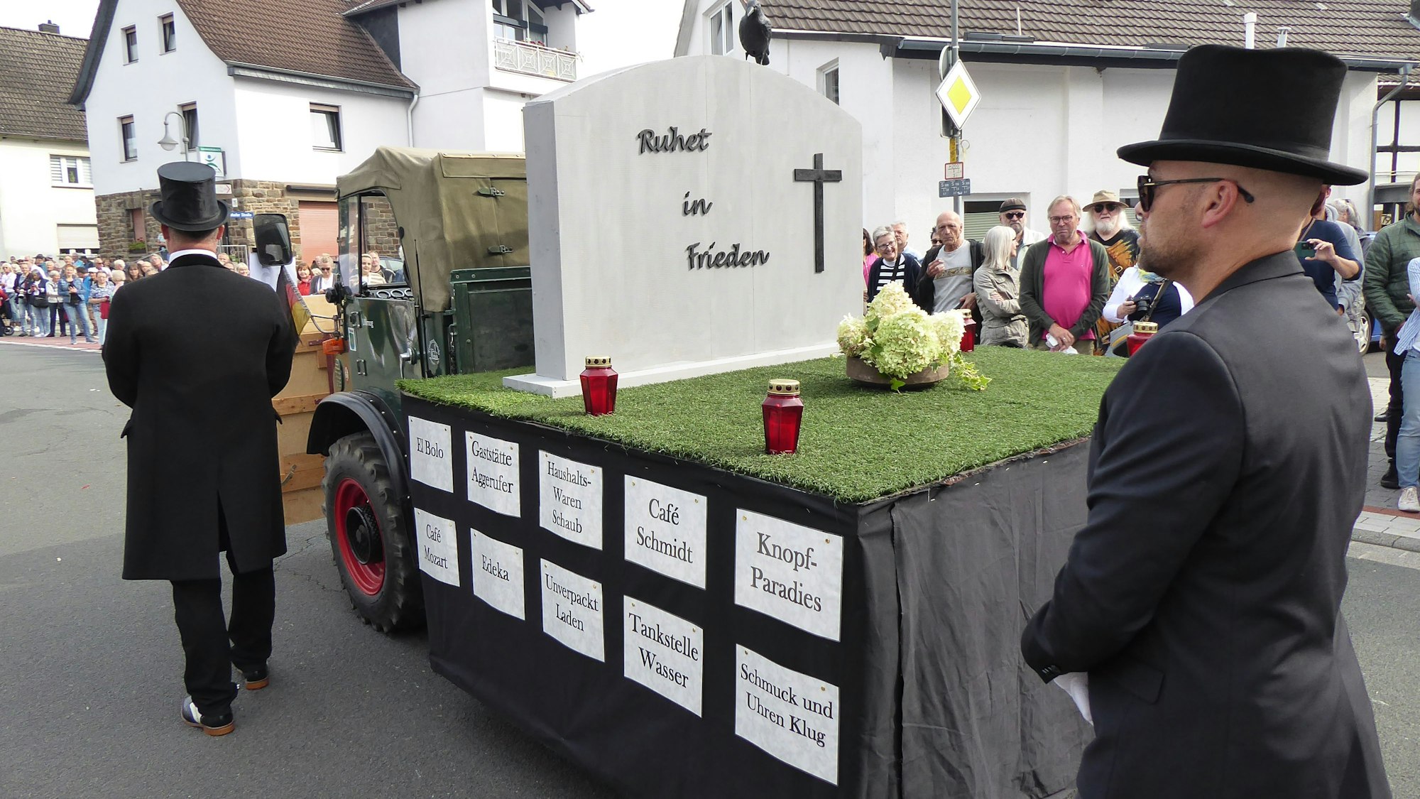 Zwei Männer in schwarzen Anzügen begleiten einen Wagen, der wie ein Friedhof aussieht. Schilder am Rand benennen die Geschäfte, die im Ort geschlossen haben.