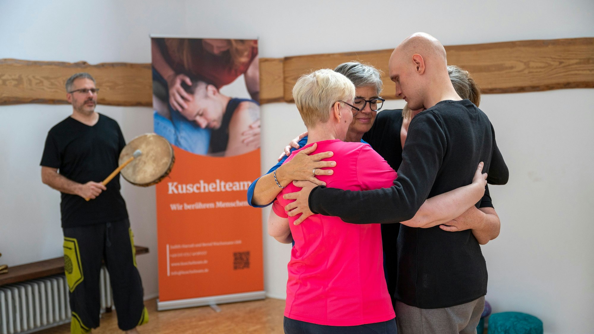 Vier Menschen umarmen sich, im Hintergrund schlägt ein Mann in eine Trommel.